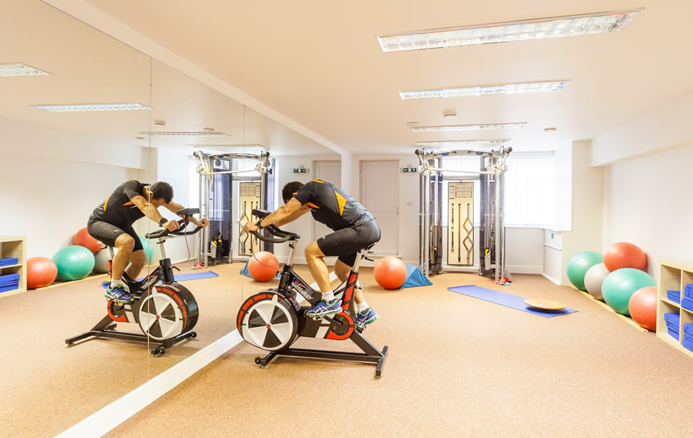 Physiotherapist using exercise bike in the Village Physiotherapy clinic, showing sports rehabilitation equipment, Pilates mats and therapy space.